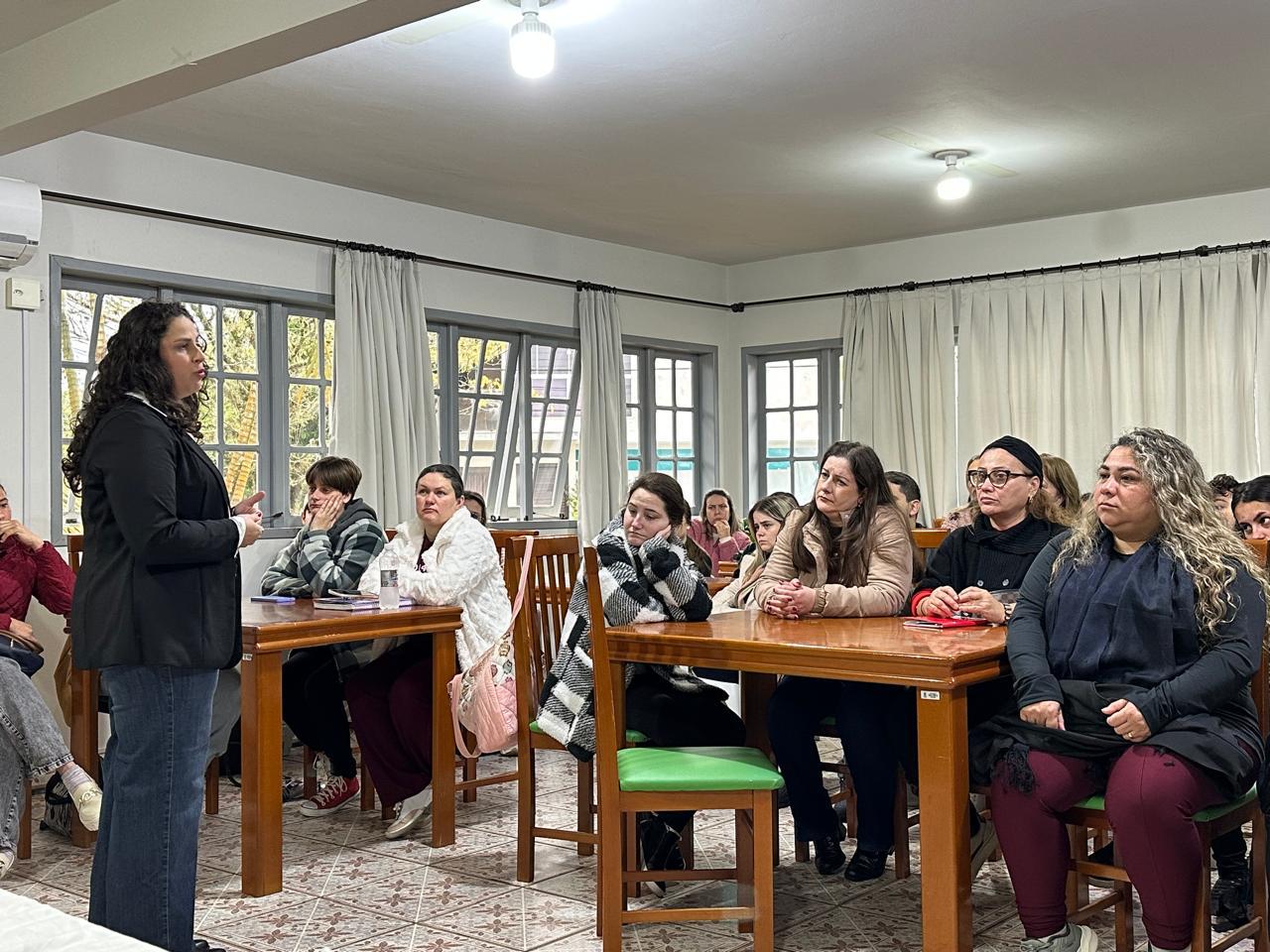 Palestrante apresenta para professoras sentadas em mesas de madeira